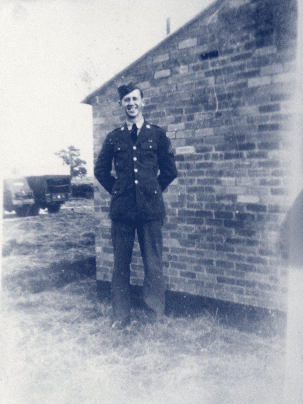 A young soldier in dress uniform standing beside a brick building, with an Army truck behind him